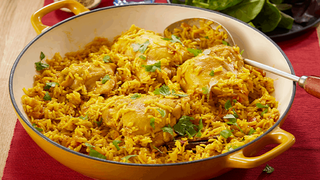 A large yellow dish filled with golden rice and tender chicken pieces, garnished with herbs. A spoon rests inside, ready to serve, with a fresh green salad in the background.