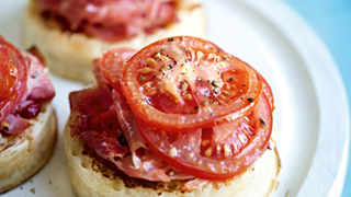 Three crumpets sit on a white plate, each topped with tomato puree, slices of wafer-thin ham and tomatoes.