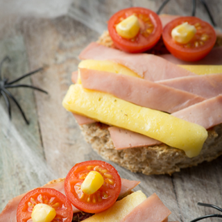 Halloween mummy face made from strips of melted cheese and turkey ham on toast, with tomatoes as eyes