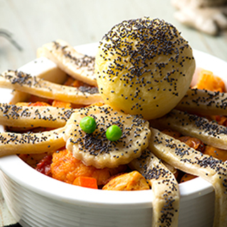 A spider made from pastry with peas as eyes sits on-top of a chicken pie, its legs resting over the bowl.