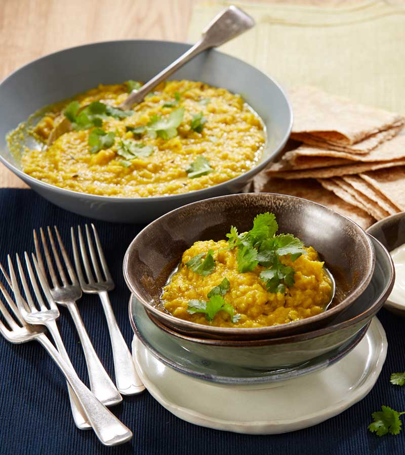Two bowls of yellow dhal, 1 small side dish and 1 main dish, topped with coriander and served with chappati and a bowl of yoghurt