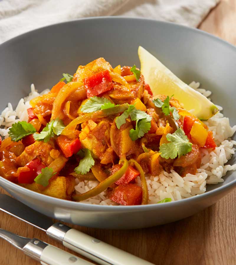 A bowl of chicken and red peppers in a spiced tomato sauce, served with a lemon wedge on a bed of rice
