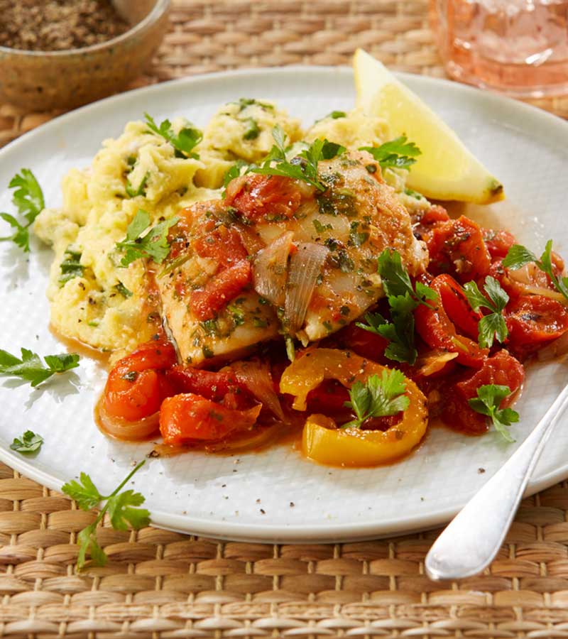 A plate of white fish fillets in tomato, pepper and thyme sauce, with a portion of okra cornmeal and a slice of lemon